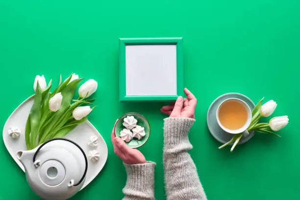 Spring celebration flat lay. Mother day, Valentine, international women day 8 or March, birthday, other special occasion. Tea pot, tea cup and white tulips, sweets. Hands hold blank frame, copy-space.