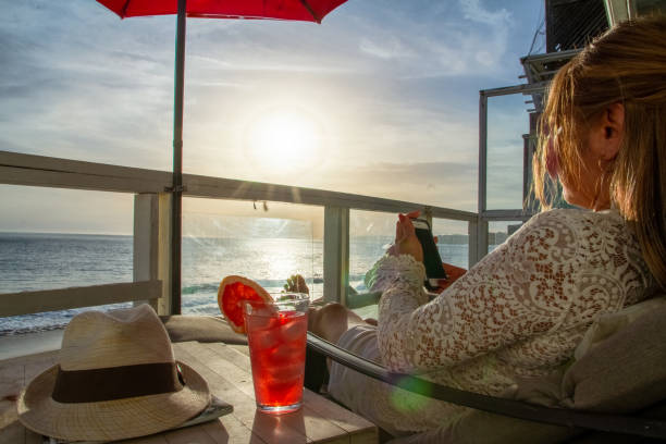海の景色を望むデッキでさわやかなドリンクを電話でテキストメッセージを送る魅力的な女性 - horizon over water malibu california usa ストックフォトと画像