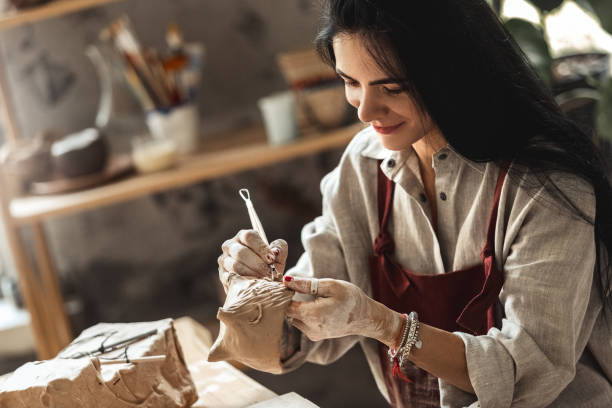 istock Craftsperson Concept. Young woman sitting at table making pottery indoors using modeling tool molding happy 1204923439