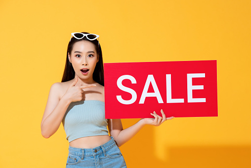 Shocked Asian woman in summer casual clothes pointing to red sale sign in hand isolated on yellow background