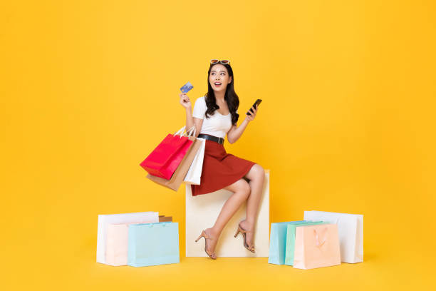 Woman carrying shopping bags with credit card and mobile phone in hands Beautiful Asian woman sitting and carrying shopping bags with credit card and mobile phone in hands on yellow background girl sitting stock pictures, royalty-free photos & images
