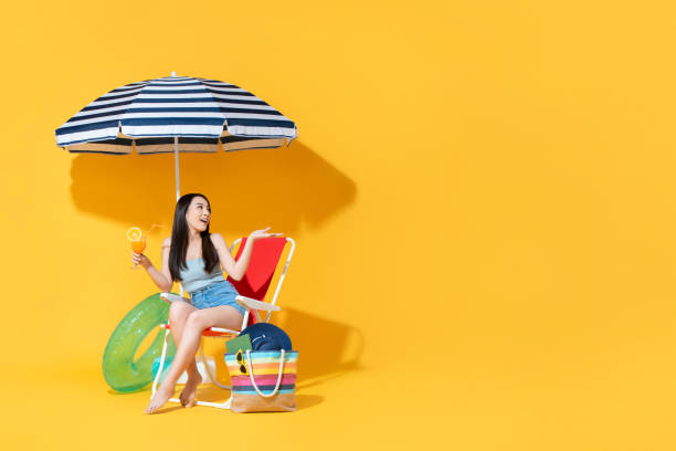 Surprised beautiful Asian woman sitting on beach chair Surprised beautiful young Asian woman sitting on beach chair doing an open palm gesture isolated on bright yellow studio background with copy space beach bag stock pictures, royalty-free photos & images