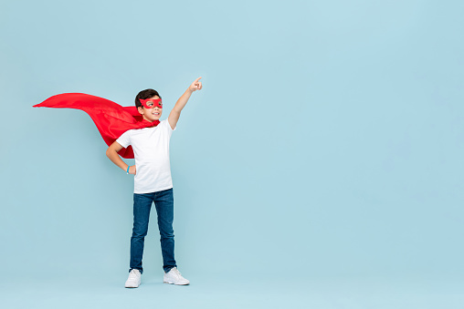 Smiling superhero boy in red mask and cape pointing hand aside isolated light blue background
