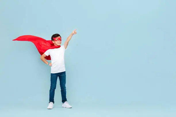 Photo of Smiling superhero boy in red mask and cape pointing hand aside