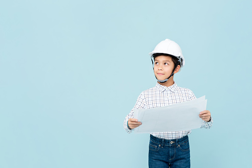 Smiling young asian boy dress like engineer with hardhat holding blueprint thinking and looking upward in light blue isolated studio backgorund