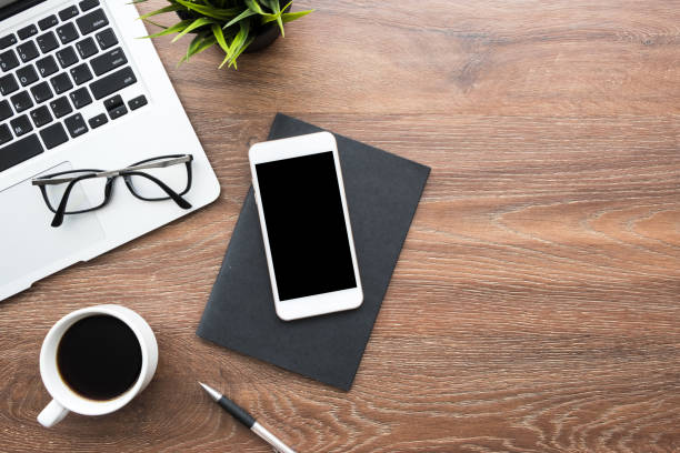 istock Wood office desk table with white smartphone with blank mock up screen, laptop computer, cup of coffee and supplies. Top view with copy space, flat lay. 1204937618