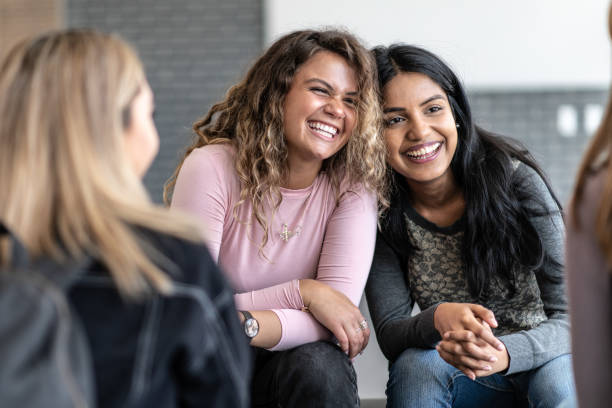 Two Girls Sharing an Embrace stock photo Two female young adults sit closely together, with their heads resting against each others, during a group therapy session.  One is of African decent and the other is of Indian decent.  Bother are dressed casually and are sitting among their multi-ethnic peers with smiles on their faces. career counseling stock pictures, royalty-free photos & images