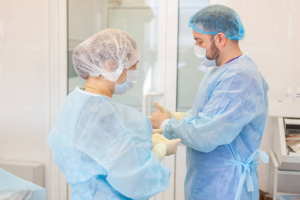 Hospital. Surgeon operates in the operating room. Profile view of a medical assistant helping a surgeon put on his gloves and medical gown in an operating room Hospital. Surgeon operates in the operating room. Profile view of a medical assistant helping a surgeon put on his gloves and medical gown in an operating room. evening gown stock pictures, royalty-free photos & images