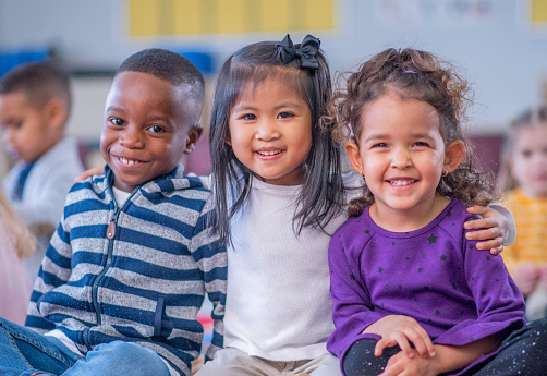 A multi-ethnic group of daycare children sit close together on the floor, with their legs crossed and their arms around one another, as they pose for a portrait.  They are smiling and enjoying their time together.