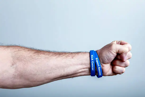 A real person, real life, senior adult man colon cancer survivor is reaching out - extending his arm and making a fist. He is wearing two cancer support themed wristbands (personally designed/"phrased"/commissioned by me - the photographer/cancer patient) embossed with the words "Kick Cancer" and "Expect Success". Part of a "Daily Living with Cancer" image series - submitted toward brief #775310942.