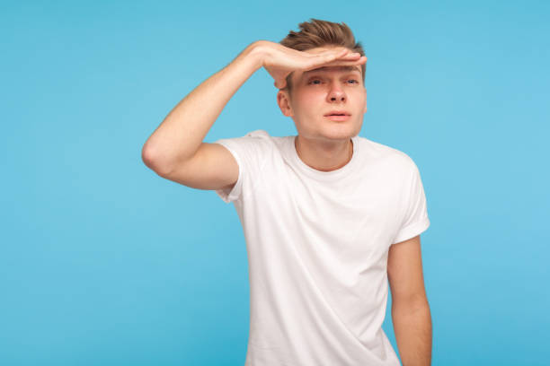 vision lointaine. verticale de l’homme curieux dans le t-shirt blanc occasionnel regardant la longue distance et retenant la main au-dessus de ses yeux - back to front rear view men people photos et images de collection