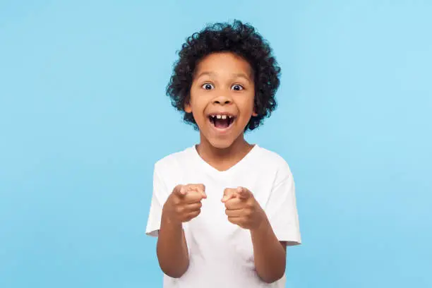 Hey you! Portrait of happy little boy with curly hair pointing finger to camera and laughing loudly with surprised face, teasing making fun of you. indoor studio shot isolated on blue background