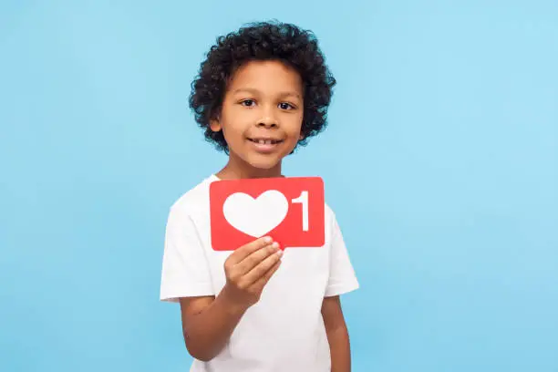 Photo of Wonderful charming little boy with curly hair holding one heart Like icon, button of social network