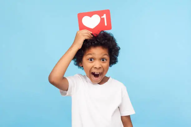 Photo of Cute surprised funny little boy with curly hair holding heart Like icon on his head and open mouth in amazement