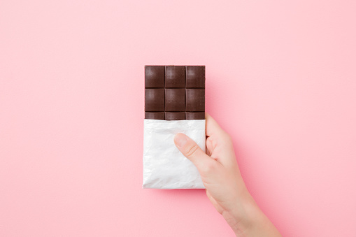 Young woman hand holding chocolate bar on light pink table background. Opened pack. Sweet snack. Closeup. Top down view.