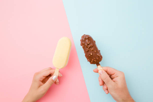 Young couple hands holding together ice creams with dark and white chocolate glaze. Closeup. Point of view shot. Light pink and blue table background. Pastel colors. Top down view. Two sides.