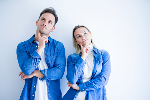 Young beautiful couple wearing denim shirt standing over isolated white background with hand on chin thinking about question, pensive expression. Smiling with thoughtful face. Doubt concept.