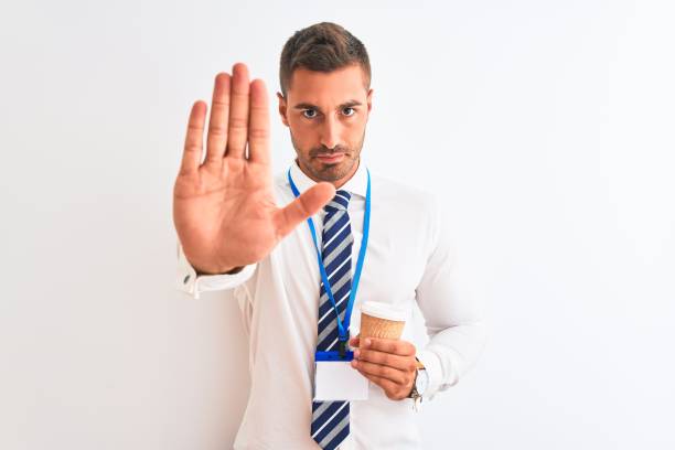 jeune homme d’affaires beau utilisant la passe d’identification et buvant le café au-dessus du fond isolé avec la main ouverte faisant le signe d’arrêt avec l’expression sérieuse et confiante, geste de défense - id card business card holding human hand photos et images de collection