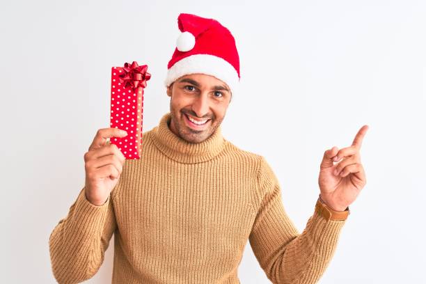 istock Young handsome man wearing christmas hat and holding gift over isolated background very happy pointing with hand and finger to the side 1204997242