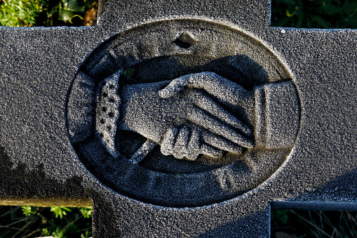 Two hands clasp each other in greeting, but they are made of stone and sprinkled with rime ice from the cold frost of a February night. Truly a frosty, cold handshake. The two hands form part of a memorial cross in a churchyard.