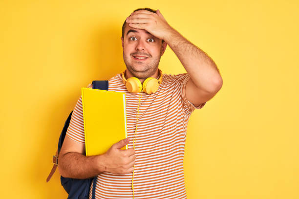 istock Student man wearing backpack headphones holding notebook over isolated yellow background stressed with hand on head, shocked with shame and surprise face, angry and frustrated. Fear and upset for mistake. 1204997754