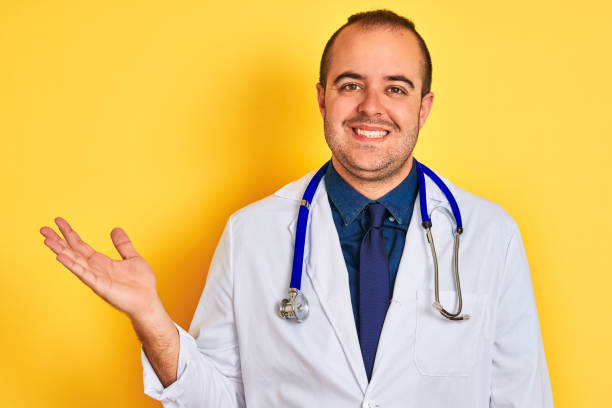 istock Young doctor man wearing coat and stethoscope standing over isolated yellow background smiling cheerful presenting and pointing with palm of hand looking at the camera. 1204998021