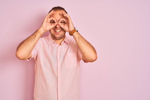 istock Young man wearing elegant shirt standing over isolated pink background doing ok gesture like binoculars sticking tongue out, eyes looking through fingers. Crazy expression. 1204998982