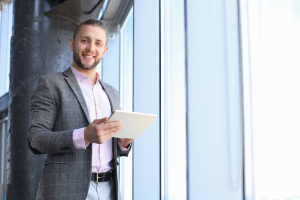 istock Handsome young business man holding digital tablet and looking at camera with smile while standing in the office. 1205000893