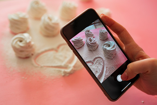Female hand take a mobile photo of sweet marshmallows desserts on pink background, food photography