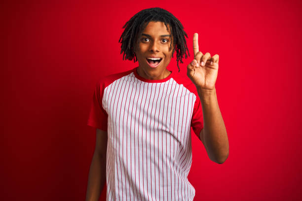 istock Afro man with dreadlocks wearing striped t-shirt standing over isolated red background pointing finger up with successful idea. Exited and happy. Number one. 1205002051