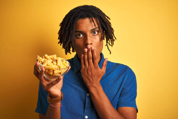 istock Afro american man with dreadlocks holding pasta macaroni over isolated yellow background cover mouth with hand shocked with shame for mistake, expression of fear, scared in silence, secret concept 1205002989