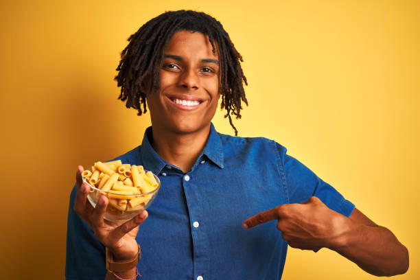 istock Afro american man with dreadlocks holding pasta macaroni over isolated yellow background with surprise face pointing finger to himself 1205003007