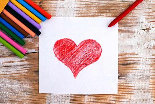 Child hand drawing Red Heart with wax crayons. Wooden background.