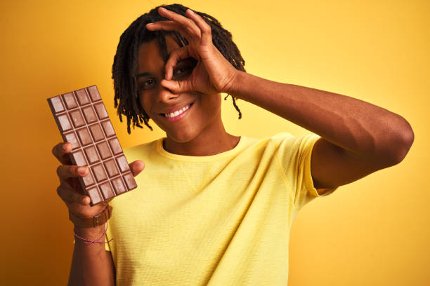 istock Afro american man with dreadlocks eating chocolate bar over isolated yellow background with happy face smiling doing ok sign with hand on eye looking through fingers 1205005132