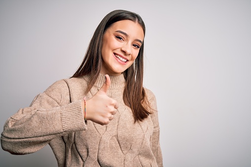 Young beautiful brunette woman wearing casual sweater standing over white background doing happy thumbs up gesture with hand. Approving expression looking at the camera showing success.