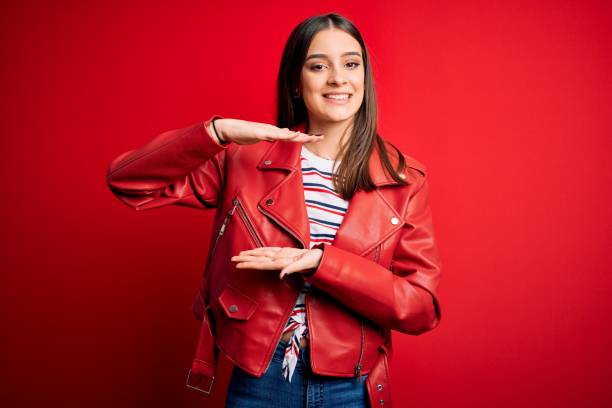 istock Young beautiful brunette woman wearing casual jacket standing over red background gesturing with hands showing big and large size sign, measure symbol. Smiling looking at the camera. Measuring concept. 1205005689