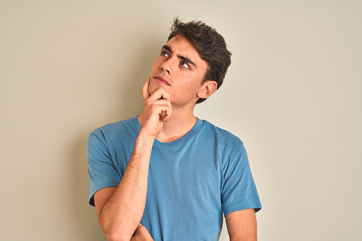 Teenager boy wearing casual t-shirt standing over isolated background with hand on chin thinking about question, pensive expression. Smiling with thoughtful face. Doubt concept.