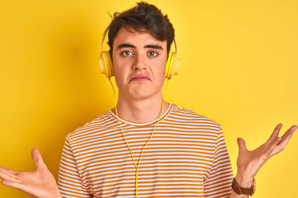 teenager boy wearing headphones over isolated yellow background clueless and confused expression with arms and hands raised. doubt concept. - head and shoulders audio imagens e fotografias de stock