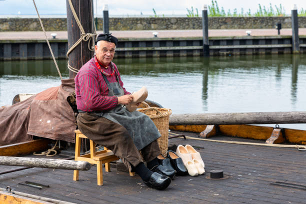 Urk, The Netherlands - September 07, 2019: Fisherman in traditional costume making wooden shoes at old historic sailing ship in Dutch harbor of Urk