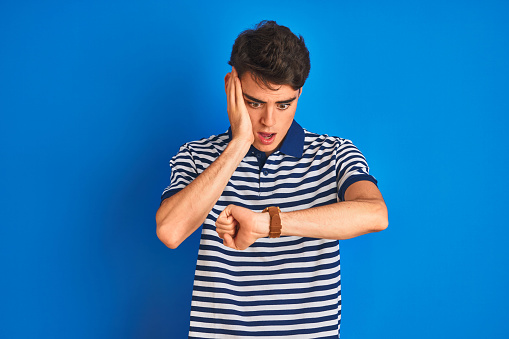 Teenager boy wearing casual t-shirt standing over blue isolated background Looking at the watch time worried, afraid of getting late