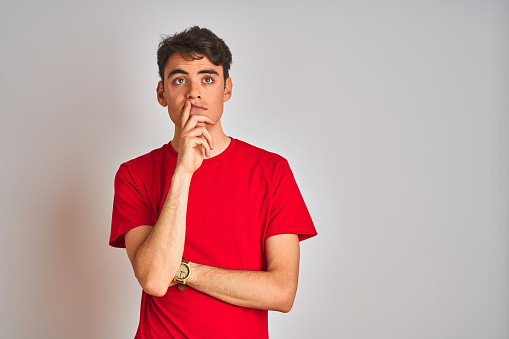 Teenager boy wearing red t-shirt over white isolated background with hand on chin thinking about question, pensive expression. Smiling with thoughtful face. Doubt concept.