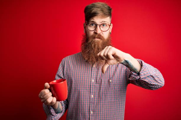 istock Redhead Irish man with beard drinking a cup of hot coffee over red background with angry face, negative sign showing dislike with thumbs down, rejection concept 1205015631