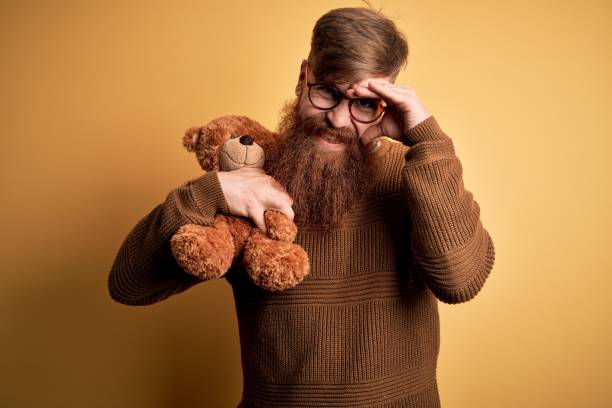 istock Redhead Irish man with beard hugging teddy bear stuffed animal over yellow isolated background stressed with hand on head, shocked with shame and surprise face, angry and frustrated. Fear and upset for mistake. 1205015804