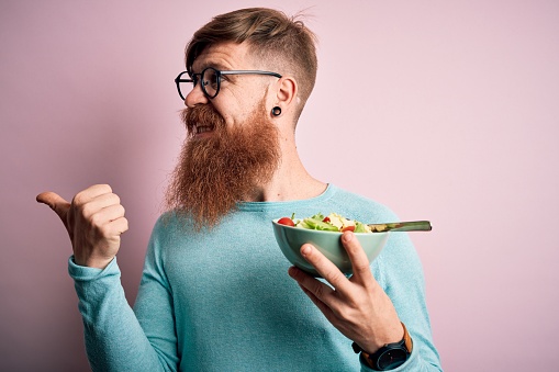 Redhead Irish healthy man with beard eating vegetarian green salad over pink background pointing and showing with thumb up to the side with happy face smiling