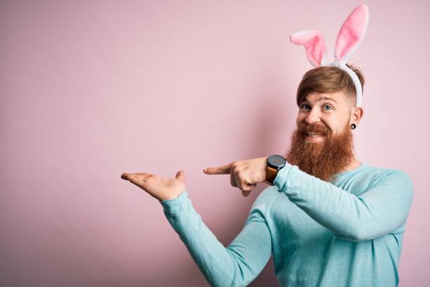 istock Hipster Irish man with beard wearing easter rabbit ears over isolated pink background amazed and smiling to the camera while presenting with hand and pointing with finger. 1205016181