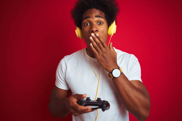 Afro gamer man playing video game using joystick headphones over isolated red background cover mouth with hand shocked with shame for mistake, expression of fear, scared in silence, secret concept