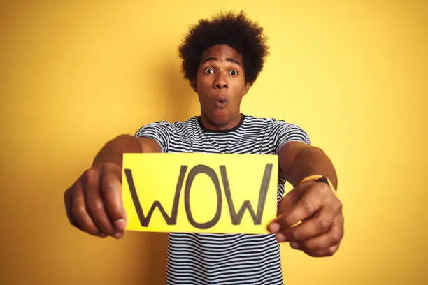 Photo of American man with afro hair holding wow banner standing over isolated yellow background scared in shock with a surprise face, afraid and excited with fear expression