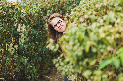 Woman hiding behind a bush - Nerd girl smiling while being behind leaves outside - Paparazzi concept image
