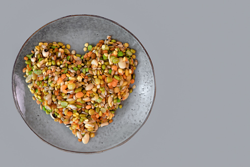 Assorted set of beans, legumes, peas, lentils laid out in the shape of a heart on a grey ceramic plate on a grey background, copy space