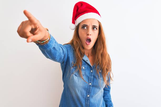 孤立した背景の上にクリスマスの帽子をかぶった若い美しい赤毛の女性は、先に驚いた指で指差し、口を開いて驚いた表情、前面に何か - front view female isolated on red happiness ストックフォトと画像